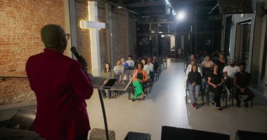 İbadet sırasında bir cemaate seslenen konuşmacı, izleyiciler sıraya dizilmiş, tuğla bir duvarda yanan haç dingin bir ruhsal atmosfer yaratıyor.