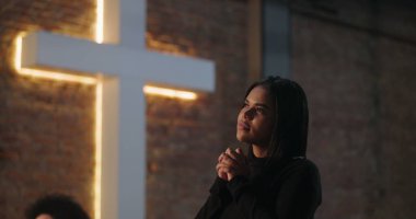 İspanyol bir kadının elleri sımsıkı bağlı, yumuşak bir ifadeyle dua edişi, evanjelist ibadet sırasında arka planda ışıklandırılmış haç, umudu, maneviyatı ve inancı sembolize ediyor.