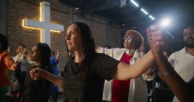 İbadet ederken el ele tutuşan, inançlarını ve bağlılıklarını ifade eden, arka planda aydınlanmış haç ruhaniliği, umudu ve yürekten birliği temsil eden bir grup insan.