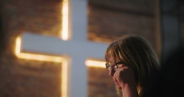 Gözyaşlarını silen kadın, derinden duygulandı, kilisedeki aydınlatılmış haçın önünde, dua sırasında duygusal deneyim, ruhani yansıma, inanç dolu an.