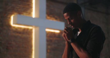 Aydınlatılmış haçın yanında ellerini sımsıkı bağlayıp dua eden adam, aşağıya bakıyor, huzur dolu ruhsal yansıma, Hristiyan inancı, ibadet ve duaya olan güven.