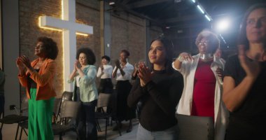 Cemaat kilise ayini sırasında el çırpıyor, arka planda haç ve tuğla duvarlar aydınlatılıyor, çeşitli topluluklar neşe ve birlik içinde.