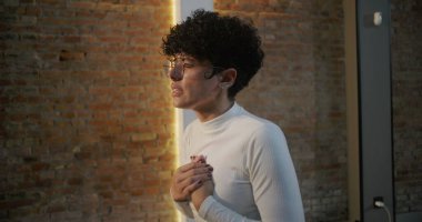 Kilisede elleri göğsüne yapışan genç bir kadın, dua ve yansıma ile meşgul, tuğla duvar ve arka planda aydınlatılmış haç, ruhani bağlılık