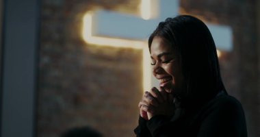 Evanjelist ibadet sırasında gülümseyen İspanyol kadının yakın çekimi, el ele tutuşmuş bağlılık, derin inanç ve ruhaniliği simgeleyen arka planda aydınlatılmış haç.