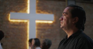 Kilise ortamında ibadet ederken derin düşüncelere dalmış bir adam, arka planda aydınlanmış haç, maneviyat, inanç ve çeşitli bir grup ortamında iç gözlem.