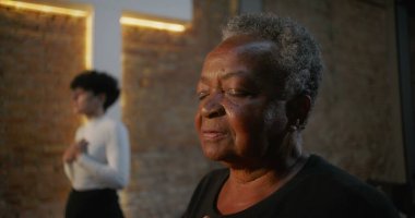 Kilisedeki yaşlı kadın ellerini göğsüne koyuyor, dua ederken gözleri kapalı, arka planda aydınlatılmış haç ve tuğla duvar, topluma tapınma ve ruhsal bağlantı.