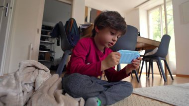 Yerde oturan genç bir çocuk, iki eliyle bir oyuncak kamera tutuyor, ev ortamında oyalanırken dikkatle odaklanıyor.