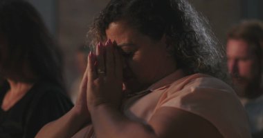 Dua eden elleri sımsıkı bağlı bir kadın, başı derin ruhani bir yansımayla eğik, huzurlu ve dingin bir bağlılık anı, ibadetle çevrili bir topluluk.