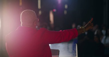 Afrika kökenli bir kadın, dini ayin sırasında tutkulu bir şekilde vaaz veriyor, cemaate, ruhani liderlere seslenirken elini kaldırıyor, ibadet edenlere inanç ve bağlılıkla rehberlik ediyor.