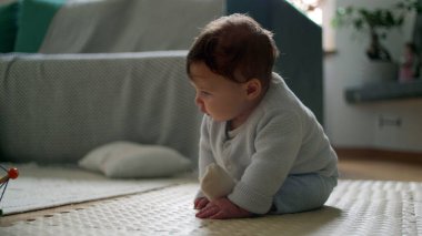 Bebek yerde oturuyor, meraklı bir ifadeyle aşağı bakıyor, rahat bir oturma odasında çevreyi keşfediyor, bağımsız, meşgul ve gözlemci olmanın ilk aşamaları..