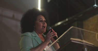 Bir kilise etkinliğinde podyumdan tutkulu bir şekilde konuşan bir kadın, seyirciyle ilgilenirken elini kaldırıyor.