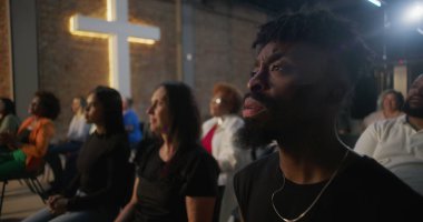 Duygusal ibadet sırasında dua eden cemaat, insanların el çırpması ve inançla el kaldırması, arka plandaki tuğla duvarda haç yakması.