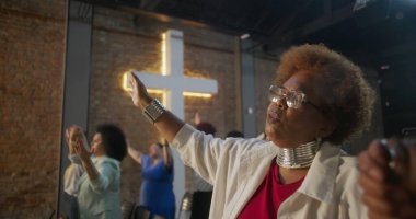 İbadet sırasında el ele tutuşan, birlik ve inanç içinde duran, arka planda toplumu ve ruhsal bağlantıyı simgeleyen haçı aydınlatan cemaat.