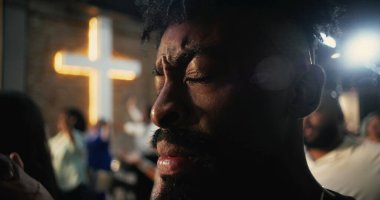 İbadet sırasında dua eden Afrikalı bir Amerikalının yakın çekimi, gözleri derin inanç ve adanmışlıkla kapalı, arka planda aydınlanan haç ruhaniliği ve umudu simgeliyor.