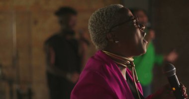 Evanjelik vaiz tutkulu bir şekilde cemaate hitap ediyor, yakın profil, gümüş saçlı Afro-soylu, gözlüklü, pembe ceketli, güçlü vaaz veren bir kadın.