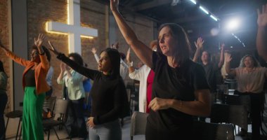 Cemaat kilise ayini sırasında el çırpıyor, arka planda haç ve tuğla duvarlar aydınlatılıyor, çeşitli topluluklar neşe ve birlik içinde.