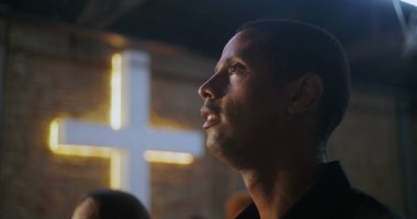 Modern bir kilise ortamında koltuğundan kalkan bir adam, arka planda tuğla bir duvarda parlayan bir haçla, ibadet sırasında odaklanmış ve ilham almış görünüyor.