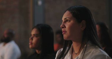 Odaklanmış ifadeye sahip bir kadın, ruhsal olay sırasında dikkatle dinliyor, dinleyicilerin arasında, huzurlu ve yansıtıcı bir inanç ve bağlantı anı yaşıyor.