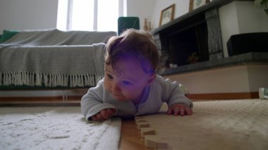 Bebek yerde yatıyor, ahşap bir bloğu yakından inceliyor, merakla eğiliyor, etkin bir şekilde çevreyi keşfediyor, erken çocukluk öğrenimi, rahat bir evde dokunsal bir keşif..