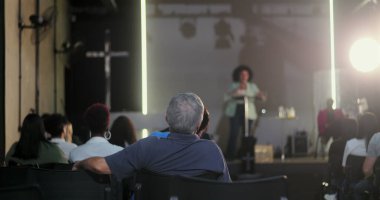 Seyirciler arasında sırtı hoparlöre dönük oturan bir adam, vaaz veren bir kadına dikkat ediyor ya da bir kilise etkinliğinde konuşma yapıyor, arka planda çapraz