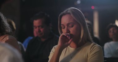 Sarışın bir kadın sessizce dua ediyor, elleri dudaklarında, gözleri kapalı, ruhsal yansıma ve huzur dolu ibadete dalmış.