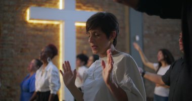 Kapalı gözlerle dua eden ve çeşitli grup ibadet ortamlarında el kaldıran kadın, arka plan, inanç, maneviyat ve toplumsal adanmışlık alanlarında aydınlatılmış haç.