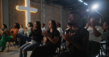 İçten bir ibadet sırasında alkış tutan cemaat, inançla ve duayla meşgul olan insanlar, tuğla bir duvarda yanan haç, ruhani bir atmosfer yaratıyor.