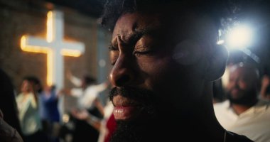 İbadet sırasında dua eden Afrikalı bir Amerikalının yakın çekimi, gözleri derin inanç ve adanmışlıkla kapalı, arka planda aydınlanan haç ruhaniliği ve umudu simgeliyor.