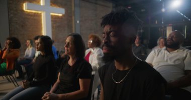 Duygusal ibadet sırasında dua eden cemaat, insanların el çırpması ve inançla el kaldırması, arka plandaki tuğla duvarda haç yakması.