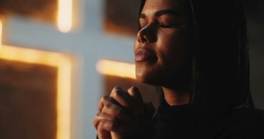 Elleri sımsıkı bağlı dua eden genç bir kadın, derin ruhsal bağ, arka planda aydınlanan haç, kilisedeki huzur dolu inanç ve bağlılığın ifadesi.