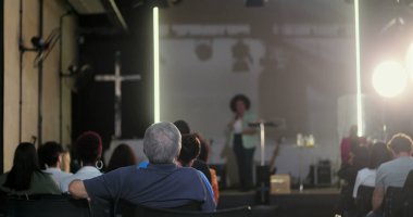 Seyirciler arasında sırtı hoparlöre dönük oturan bir adam, vaaz veren bir kadına dikkat ediyor ya da bir kilise etkinliğinde konuşma yapıyor, arka planda çapraz