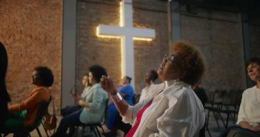Kilise ayini sırasında şarkı söyleyen ve ibadet eden çeşitli cemaatler, arka planda aydınlatılmış haç ve tuğla duvar, maneviyat ve inancı ifade ediyor.