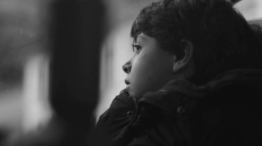 Hareket halindeki bir otobüste pencereden dışarı bakan genç bir çocuk, sessizce düşüncelere dalmış, dışarıdaki dünyayı sakin bir ifadeyle gözlemliyor, siyah ve beyaz.