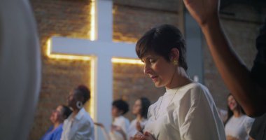 Kapalı gözlerle dua eden ve çeşitli grup ibadet ortamlarında el kaldıran kadın, arka plan, inanç, maneviyat ve toplumsal adanmışlık alanlarında aydınlatılmış haç.