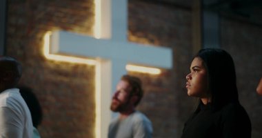 Siyahlı genç bir kadın, ciddi bir ifadeyle, arka planda aydınlanmış haç, Hıristiyan inancını, tefekküre ve çeşitli cemaatleri vurgulayan ruhani bir toplantı.