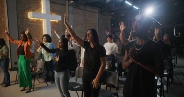 Cemaat kilise ayini sırasında el çırpıyor, arka planda haç ve tuğla duvarlar aydınlatılıyor, çeşitli topluluklar neşe ve birlik içinde.