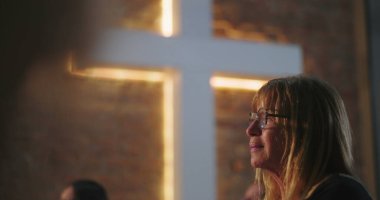 Kilisede aydınlatılmış haç önünde yaşlı bir kadının portresi, dua sırasında duygusal deneyim, ruhani yansıma, inanç dolu bir an.