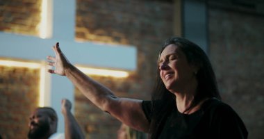 İbadet eden bir kadın, dua ederken gözlerini kapatarak elini kaldırıyor, duygusal bağ kuruyor, Hıristiyan ruhaniliği, tuğla duvarda aydınlatılmış haç, yakınlardaki çeşitli cemaatler.