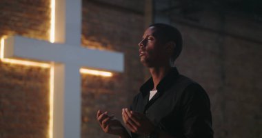 Gözler kapalı dua eden bir adam, dua ederken elleri nazikçe kaldırılmış büyük bir haçın yanında, huzurlu bir yansıma, ruhsal bağlantı ve inanç.