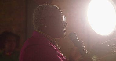 İbadet töreni sırasında mikrofona konuşan Afrikalı Amerikalı vaiz, güçlü Hıristiyan topluluklarında liderlik, inanç ve bağlılığın altını çizen arka plandaki siluet.