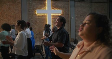 İçten ibadete katılan bireyler ellerini kaldırmış, arka planda haç aydınlatmış, kilise ayini sırasında sadakat, birlik ve inanç ifadeleri sergiliyorlar.