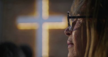 İbadet sırasında huzur içinde dua eden yaşlı kadın, arka plan, inanç, maneviyat ve içgözlem konularında aydınlatılmış haç,