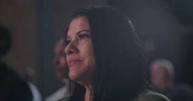 Orta yaşlı bir kadın, uzun siyah saçlı, yan görünüşlü, dikkatli, odaklanmış, yüzünden gözyaşları akıyor, bir grup ortamında derin duygusal konsantrasyon gösteriyor.