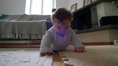 Bebek yerde yatıyor, ahşap bir bloğu yakından inceliyor, merakla eğiliyor, etkin bir şekilde çevreyi keşfediyor, erken çocukluk öğrenimi, rahat bir evde dokunsal bir keşif..