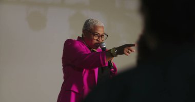 Sözcü tutkulu bir şekilde dinleyicilere hitap ediyor, mikrofonu tutuyor, vurgu için elini işaret ediyor, güçlü vaaz dağıtımı, çeşitli cemaatler dikkatlice dinliyor, ruhani olay