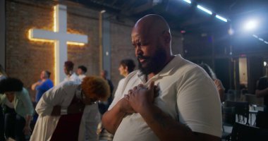 Kilisedeki sandalyeden kalkan, ellerini göğsüne koyan, dua ederken gözlerini kapayan, arka planda ışık saçan, topluma tapınan ve ruhani yansıması olan bir adam.