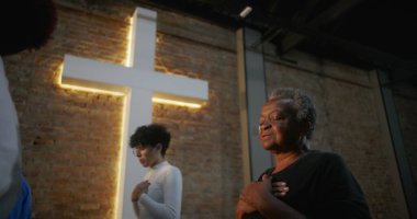 Kilisedeki yaşlı kadın ellerini göğsüne koyuyor, dua ederken gözleri kapalı, arka planda aydınlatılmış haç ve tuğla duvar, topluma tapınma ve ruhsal bağlantı.