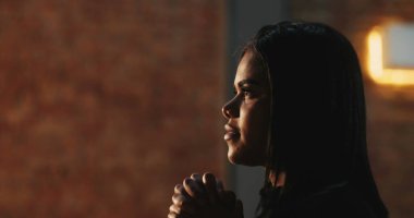 Elleri sımsıkı bağlı dua eden genç bir kadının yan profili, inancın huzurlu ifadesi, yumuşak bir ışık, arka planda dokulu tuğla bir duvar, ruhani bir bağlılık.