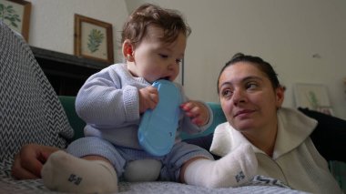 Bebek koltukta oturup mavi nesneyi çiğniyor, anne sevgiyle izliyor, kapalı alanda oyun oynuyor, bebek keşif yapıyor, aile bağı kuruyor, rahatlamış ev ortamları, çocuk gelişimi..