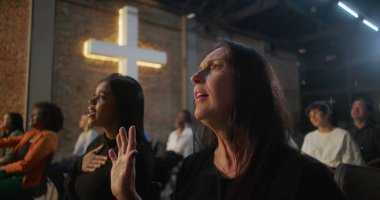 Kilise ayini sırasında dua eden kadınlar, ibadet eden biri elini kaldırırken, diğeri bağlılık, haç ayini ve arka plandaki cemaat ayini için göğsünü tutuyor.